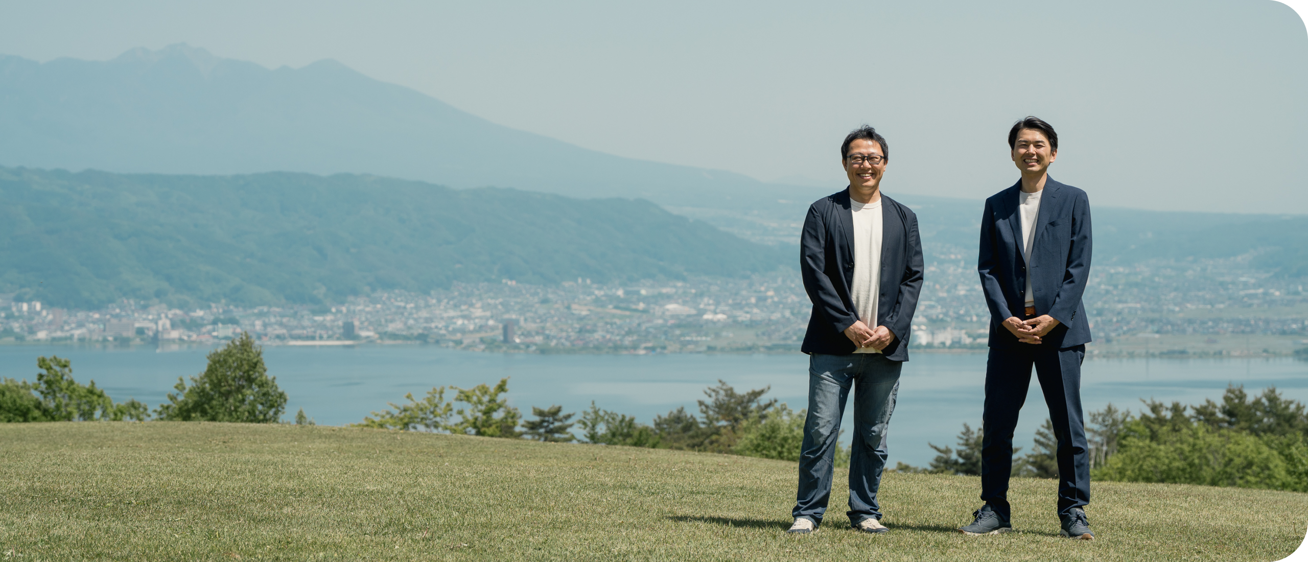 諏訪湖と山々を望む丘に立つ合同会社なまけもののメンバー